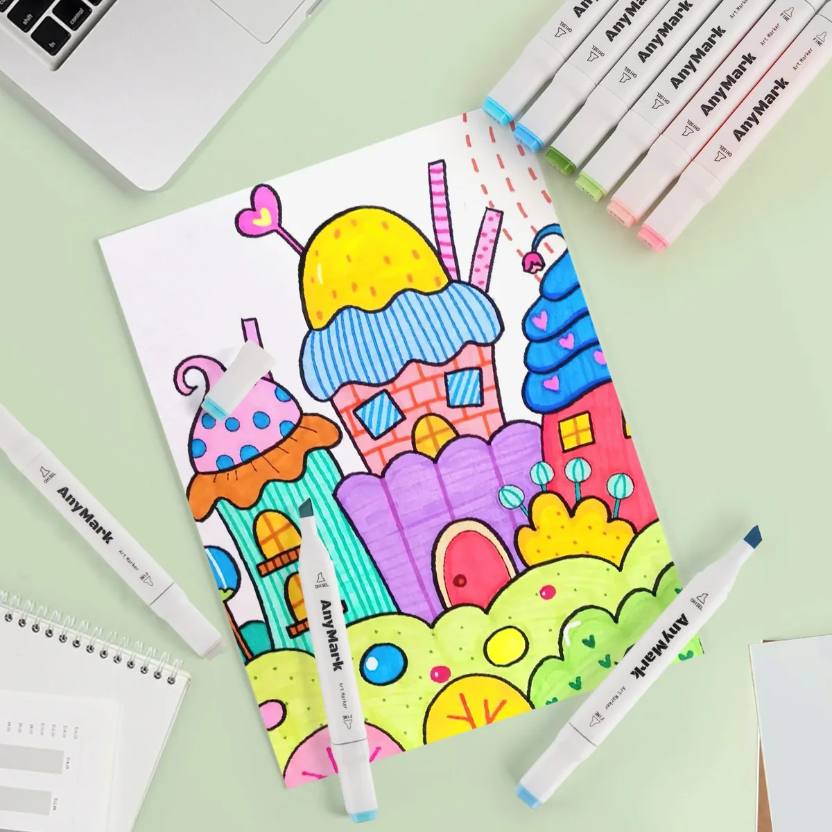 A hand-drawn page of cupcake houses colored in bright yellow, blue, pink, purple and green with AnyMark markers lying around it